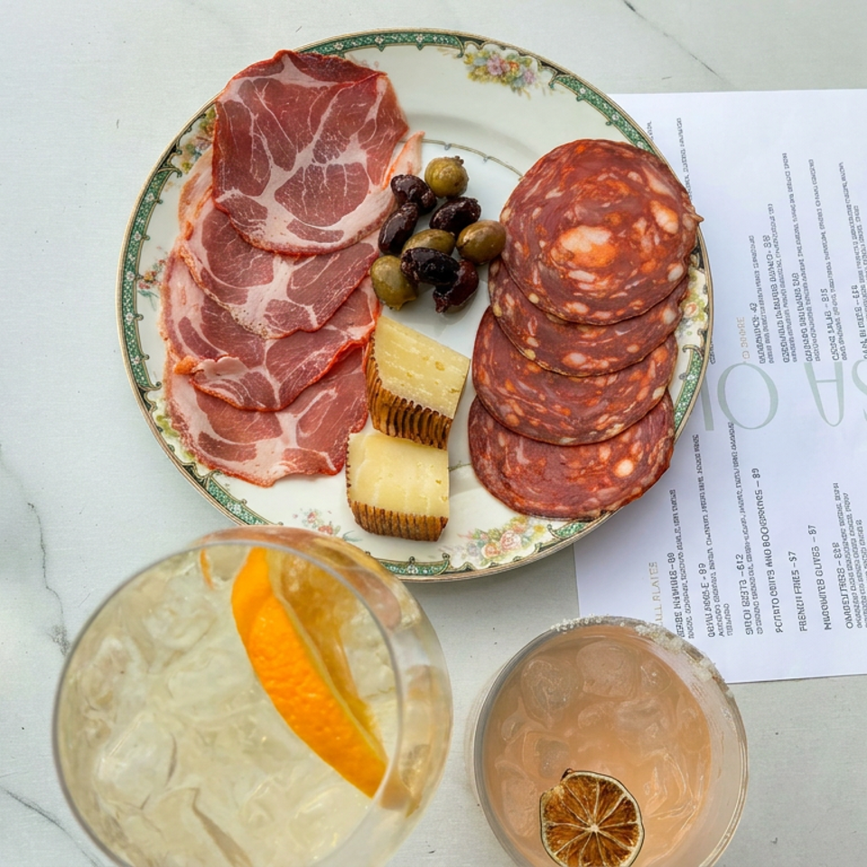 Charcuterie plate with cured meats, cheese, and olives beside two cocktails on a marble table.