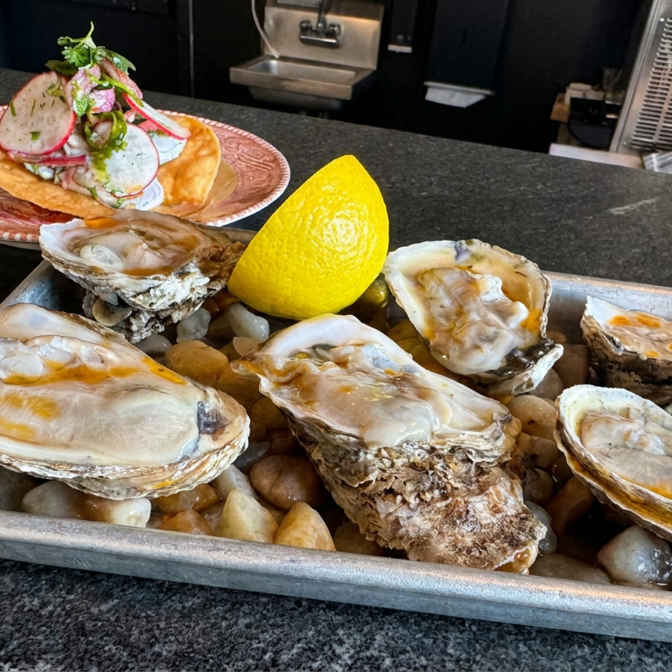 Fresh oysters on ice with a lemon wedge, accompanied by a garnished tostada on a restaurant counter.